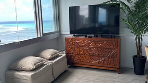 A modern living room with a large window overlooking the ocean, a flat-screen TV on a wooden cabinet, a beige sofa, and a potted plant. - Cozy studio in Waikiki  across the street from Waikiki Beach (Honolulu)