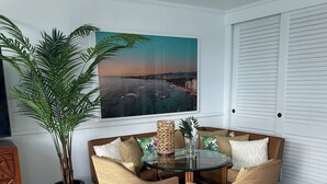 A dining area with a glass table and wicker chairs, a potted plant, and a large cityscape painting on the wall. - Cozy studio in Waikiki  across the street from Waikiki Beach (Honolulu)