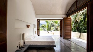 A modern bedroom with a bed, bedside table, lamp, and a view of palm trees through an open door.