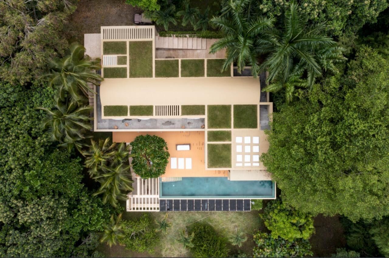 An aerial view of a modern house with a swimming pool, surrounded by lush greenery and palm trees.