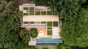 An aerial view of a modern house with a swimming pool, surrounded by lush greenery and palm trees.