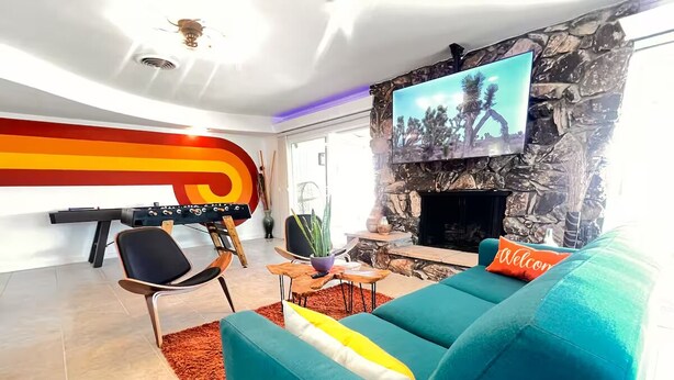 A modern living room with a teal sofa, a black chair, a TV mounted on a stone wall, and a fireplace. - Atomic Arcade: Mid-Century Getaway with Games & Pool (Yucca Valley)