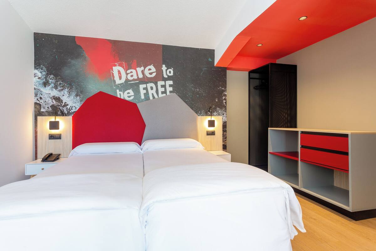 A modern hotel room with a large bed, a red accent wall, and a wall-mounted TV displaying "Date to be FREE".