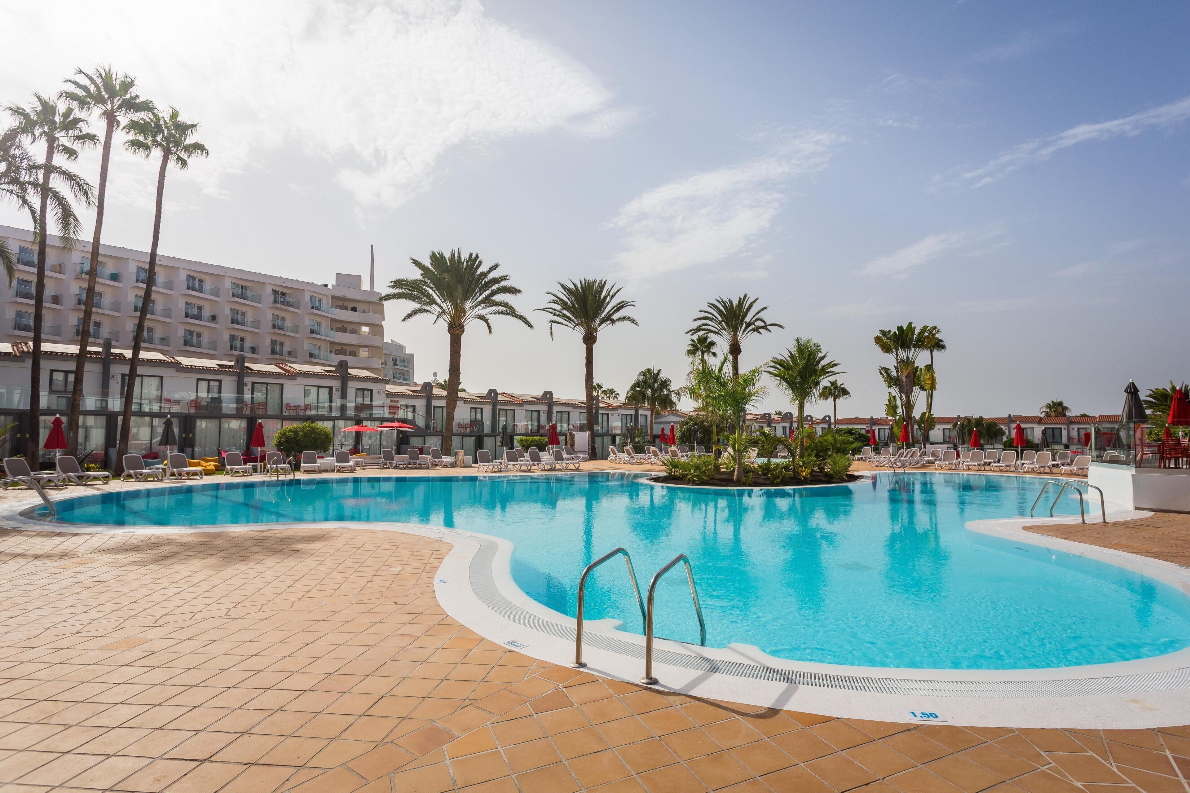 A swimming pool with lounge chairs and palm trees, surrounded by a building with multiple floors and balconies.