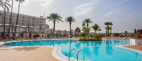 A swimming pool with lounge chairs and palm trees, surrounded by a building with multiple floors and balconies.