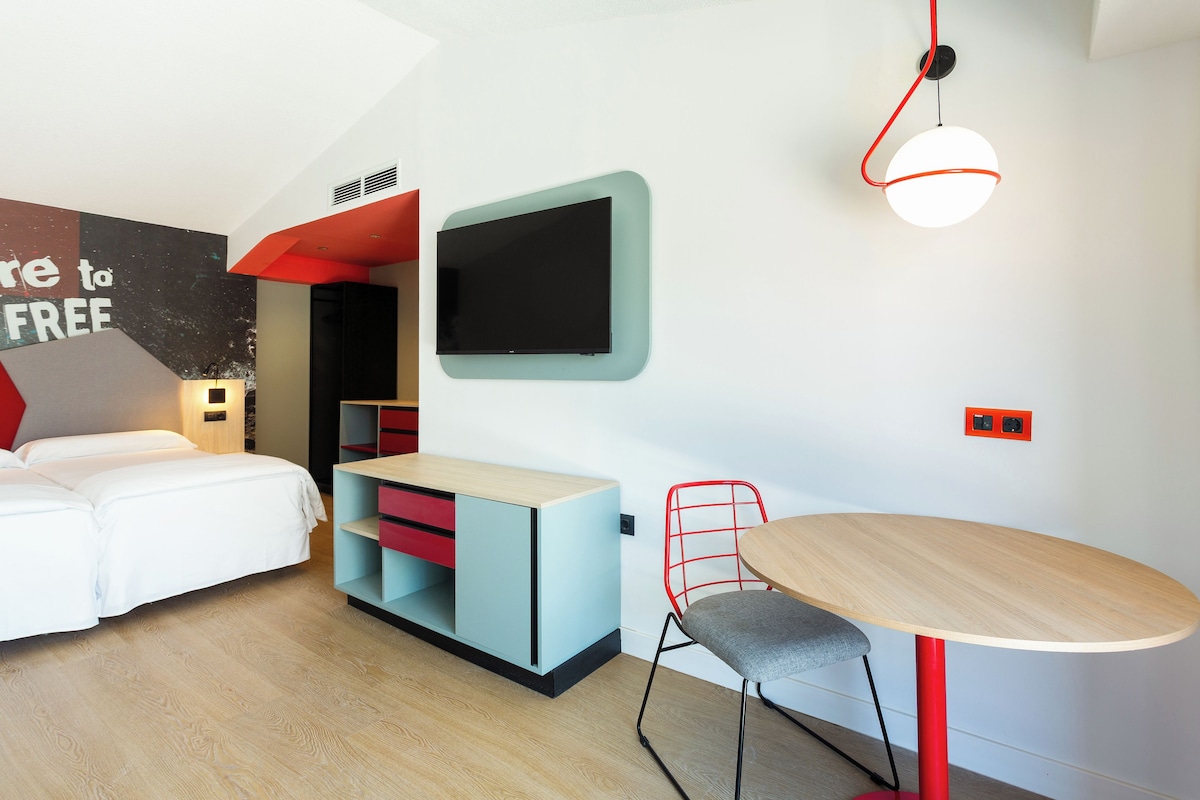 A modern hotel room with a bed, a desk with a chair, a TV mounted on the wall, and a unique red and white pendant light.