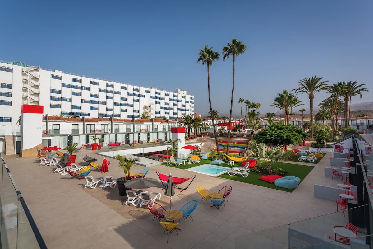 A hotel courtyard with outdoor seating, a pool, palm trees, and a modern building with multiple floors.