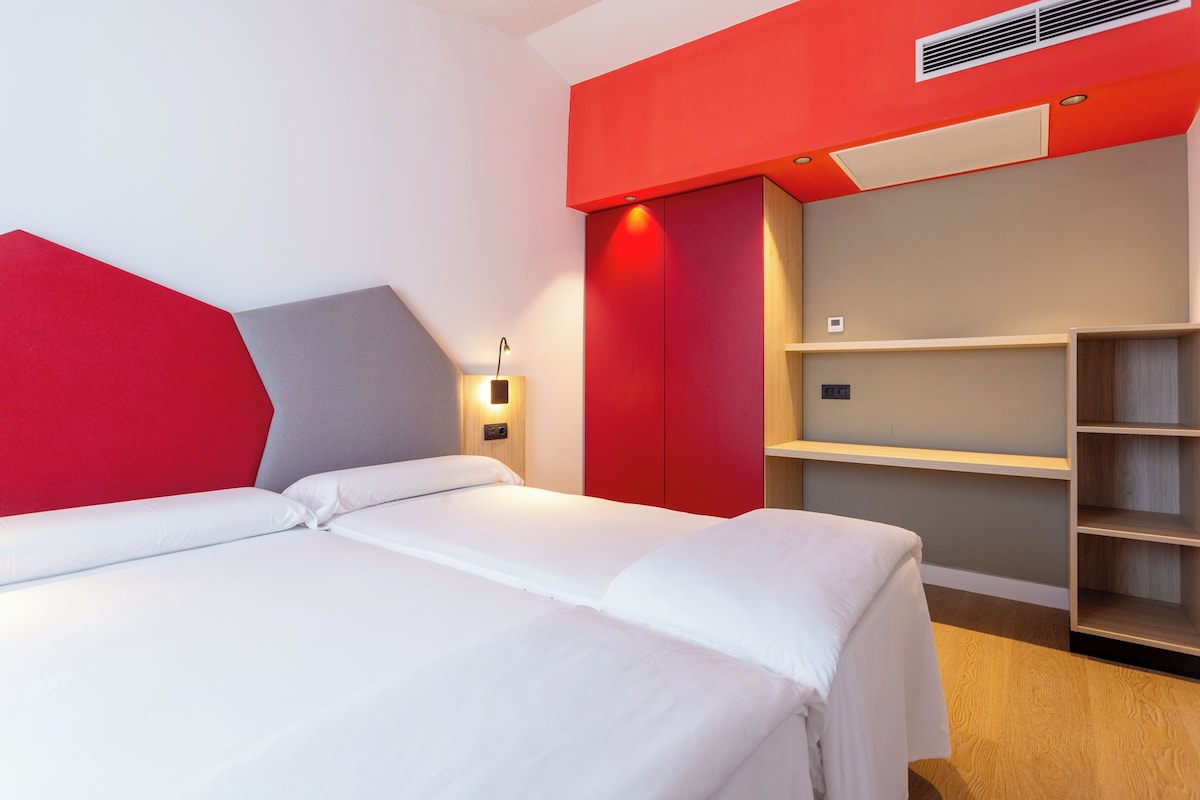 A modern hotel room with a large bed, a red accent wall, and built-in shelving.