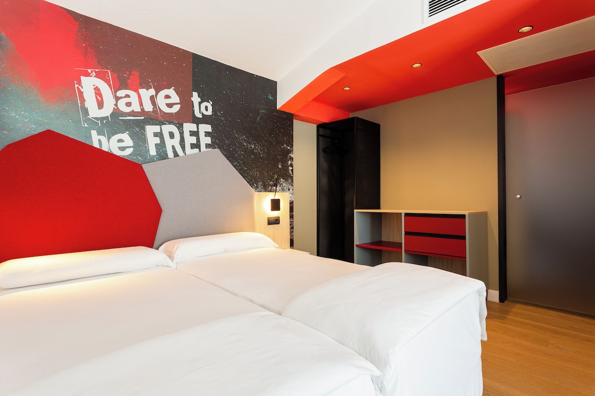 A modern hotel room with a large bed, a red accent wall, and a motivational poster.