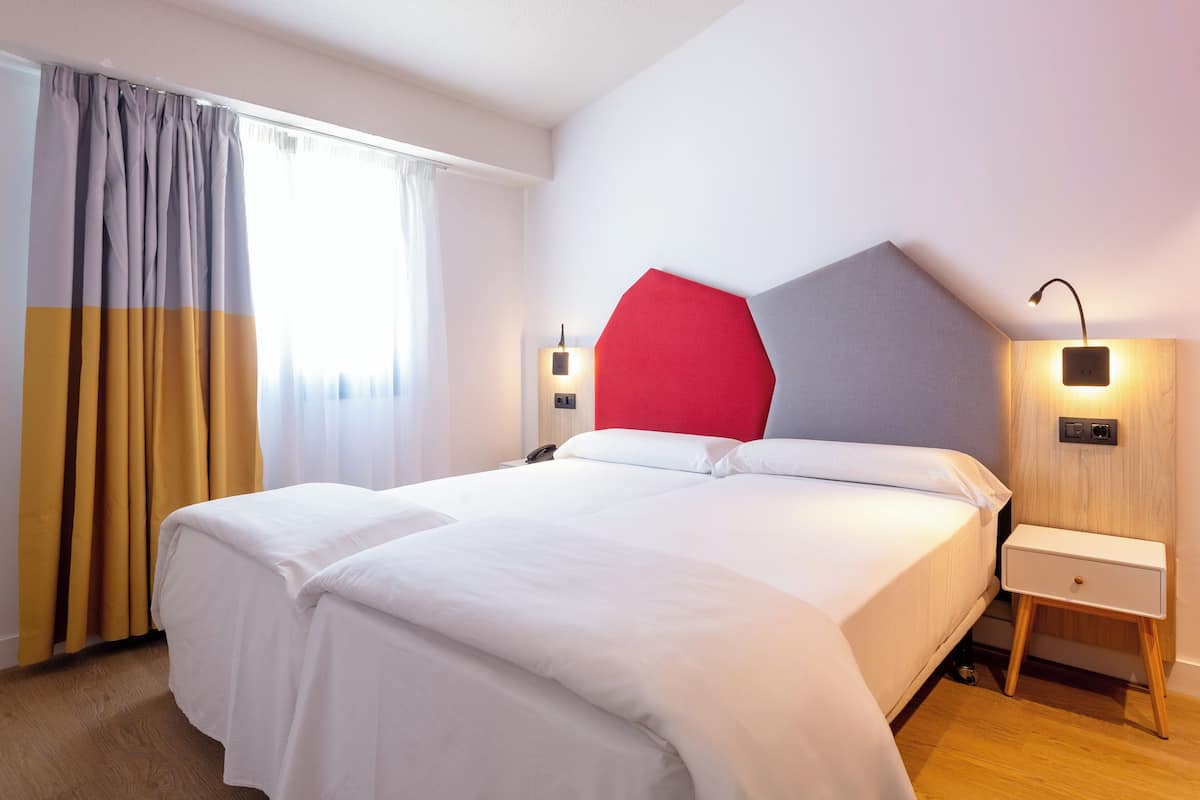 A neatly made bed with white bedding, a red accent pillow, and a wooden nightstand with a lamp.