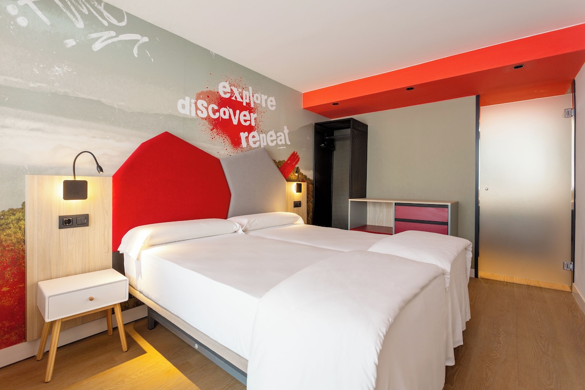 A modern hotel room with a large bed, bedside tables, a wardrobe, and a wall with a motivational quote.