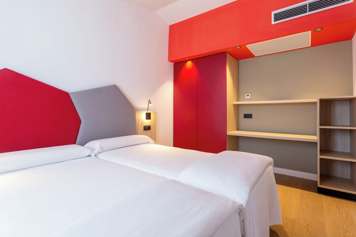 A modern hotel room with a large bed, a red accent wall, and built-in shelving.