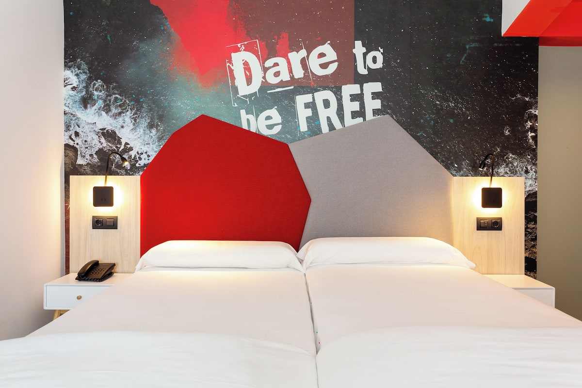 A modern hotel room with a large bed, a red accent wall, and a motivational poster.