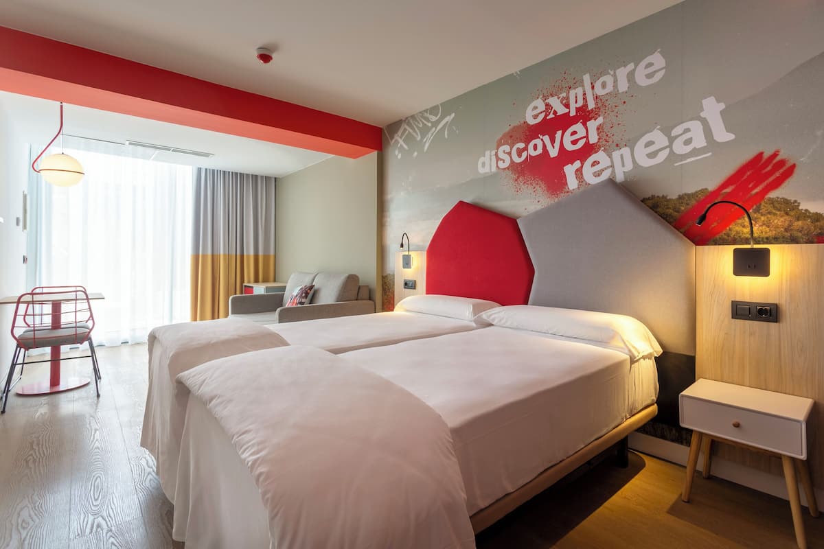 A hotel room with two beds, a red accent wall, a wall-mounted lamp, and a small bedside table.