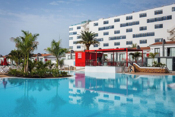Una piscina con sillones de descanso y palmeras, rodeada por un edificio con varias ventanas y una estructura roja.