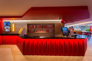 A reception area with a red counter, a digital kiosk, and a wall-mounted TV. - Sholeo Lodges Maspalomas (San Bartolomé de Tirajana)