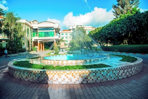 Una fuente circular con borde de azulejos, rodeada de un jardín ajardinado y un edificio con múltiples ventanas y balcones.