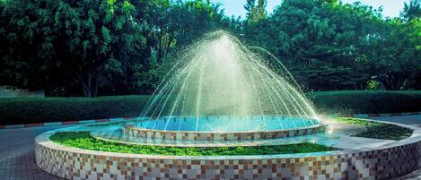 A circular fountain with water jets, surrounded by a tiled walkway and greenery.