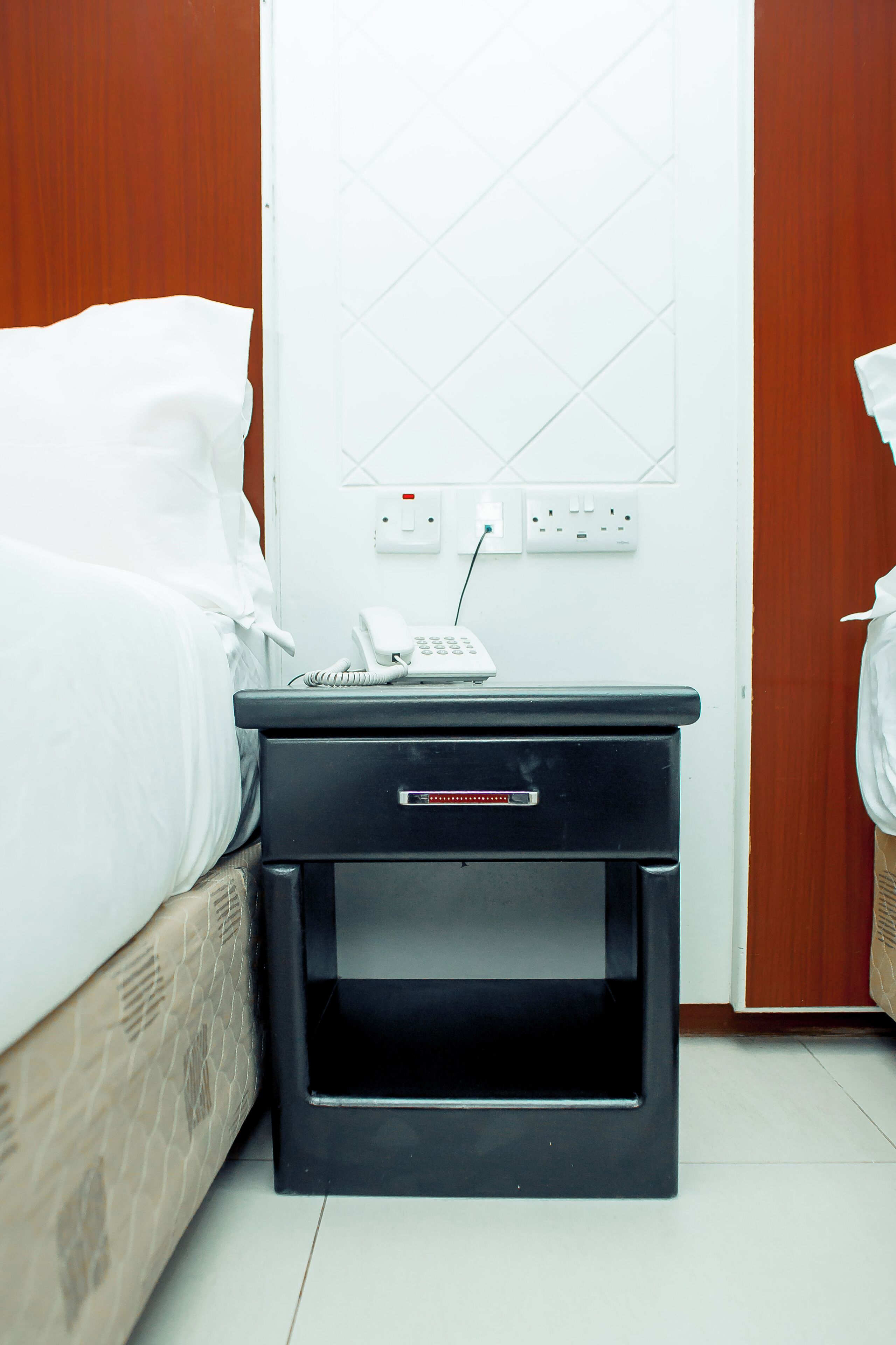 A hotel room with a bed, a bedside table with a phone and a notepad, and a wall socket.