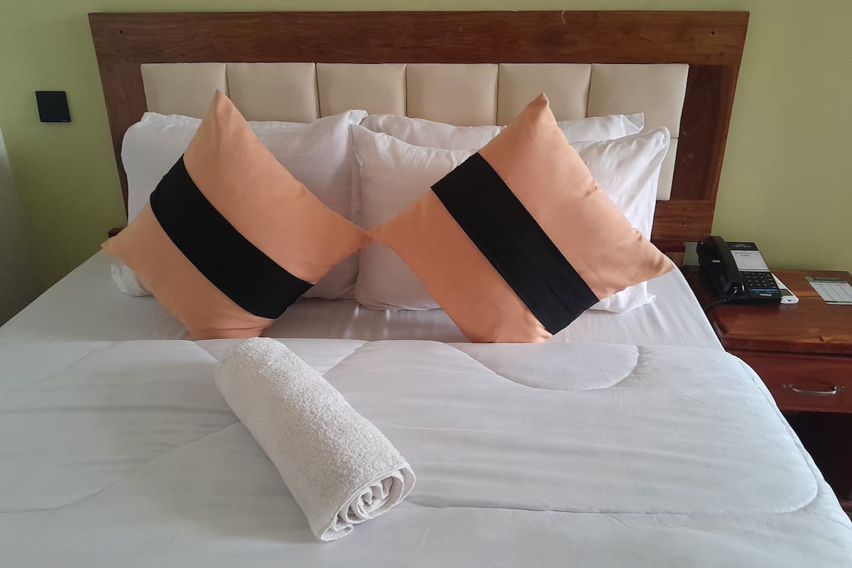 Una cama bien tendida con sábanas blancas, dos almohadas color melocotón con rayas negras y una toalla blanca enrollada.