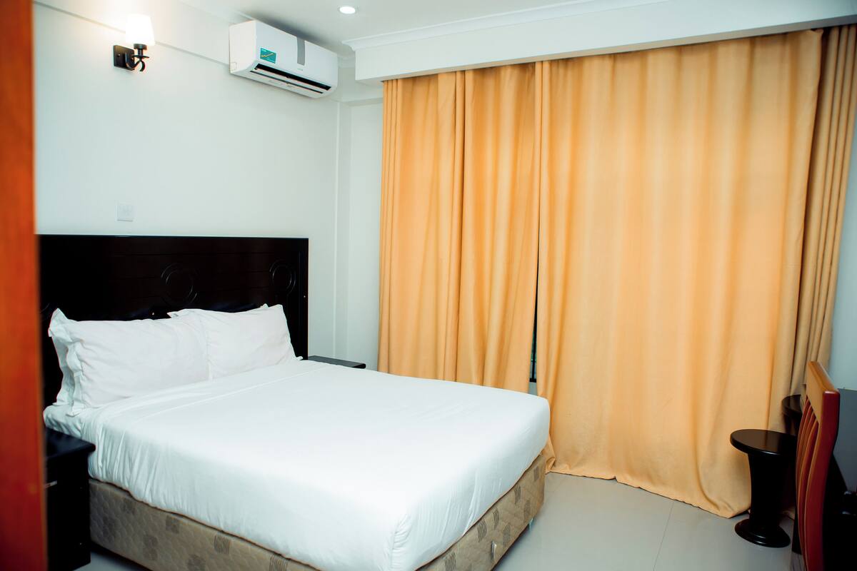 Habitación de hotel con cama, aire acondicionado, cortina, una mesita y una silla.
