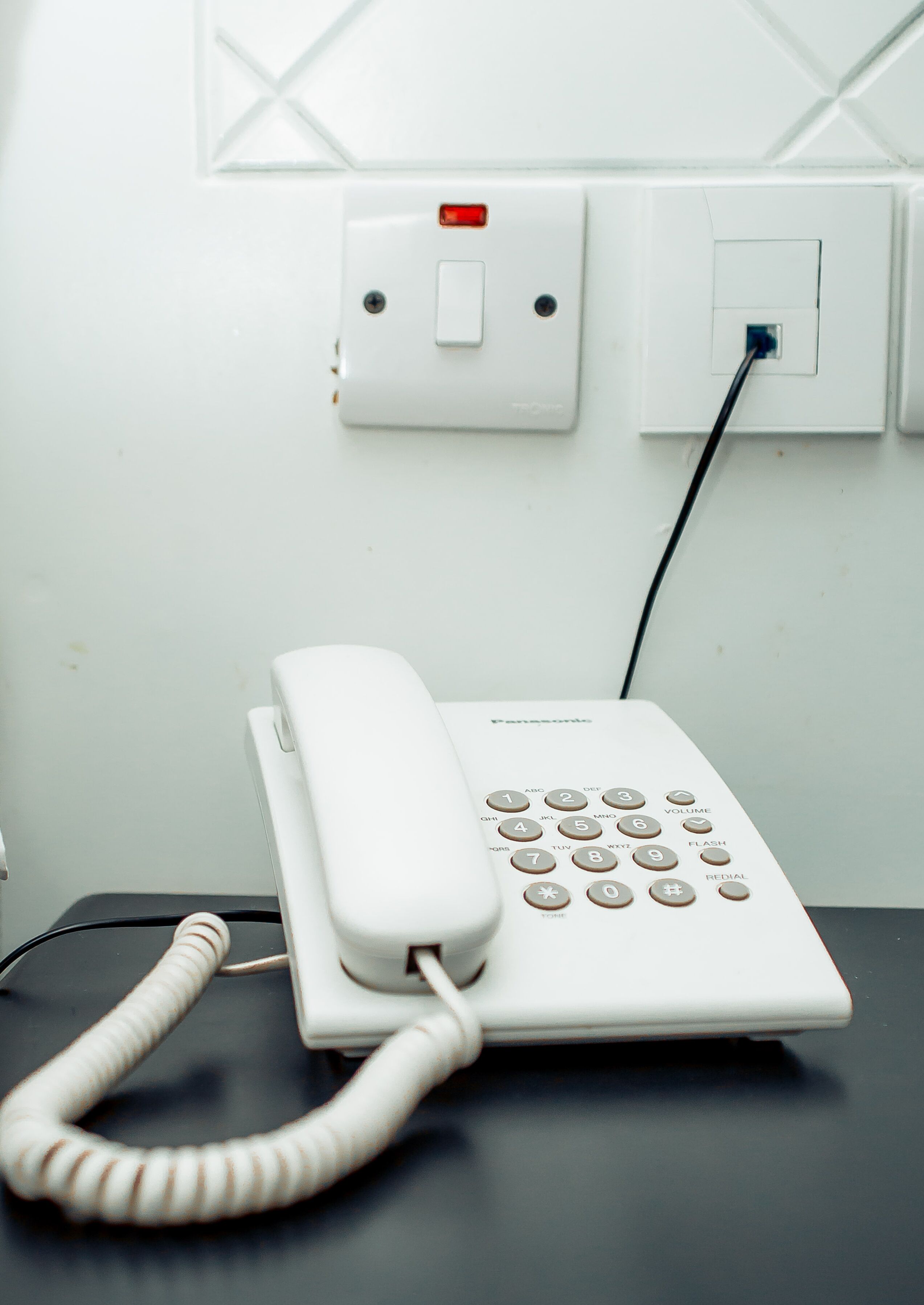 Un teléfono blanco de Panasonic sobre un escritorio con cable, conectado a un enchufe de pared y una toma de corriente.