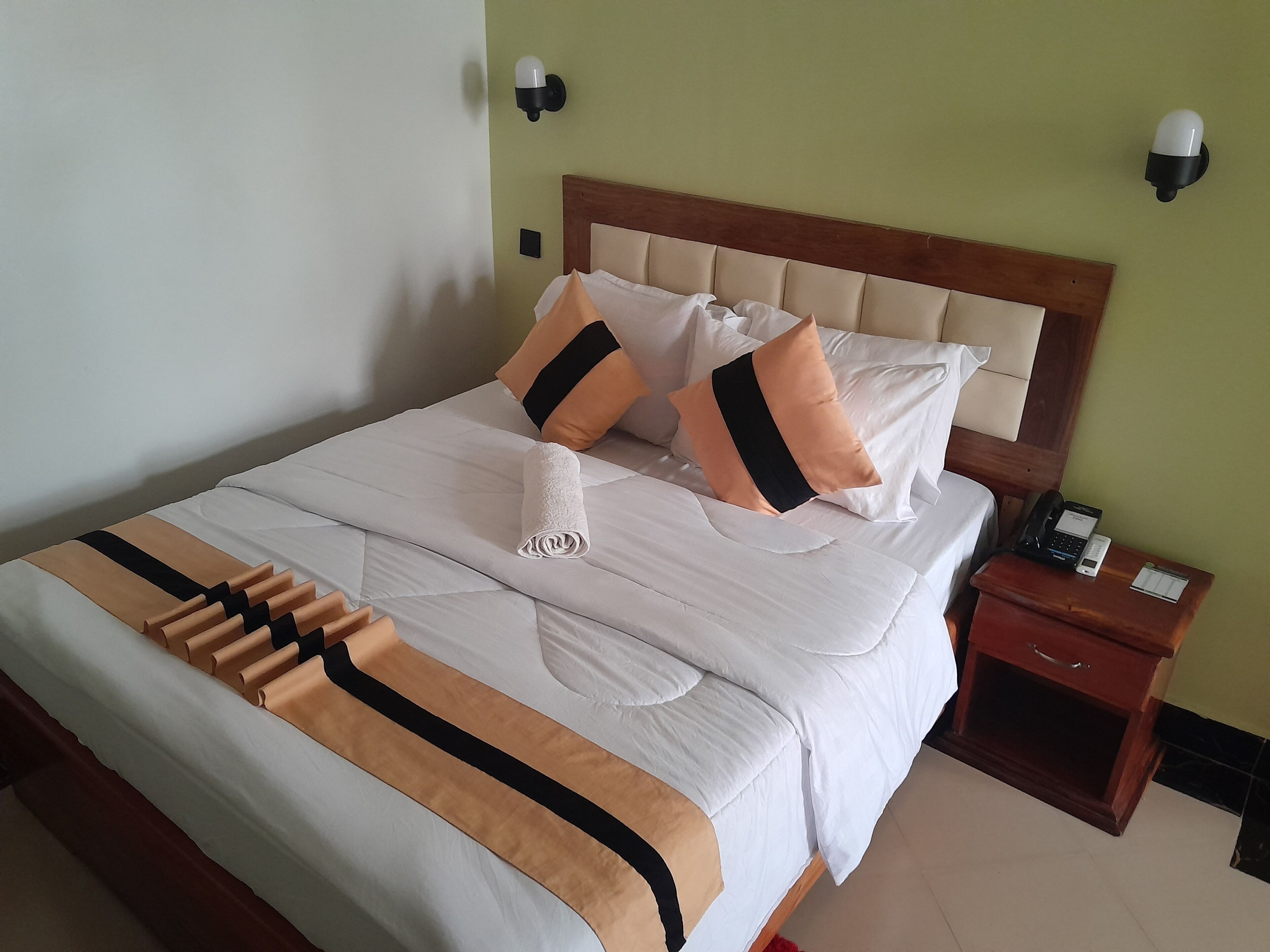 A neatly made bed with a white and brown striped bedspread, two pillows, and a folded towel. There is a wooden nightstand with a phone and a small drawer.