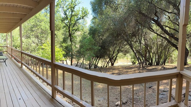 A covered outdoor deck with a railing, a table, and chairs, overlooking a wooded area.