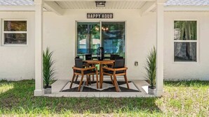 A house with a covered porch, a 'Happy Hour' sign, a dining table with chairs, and potted plants.