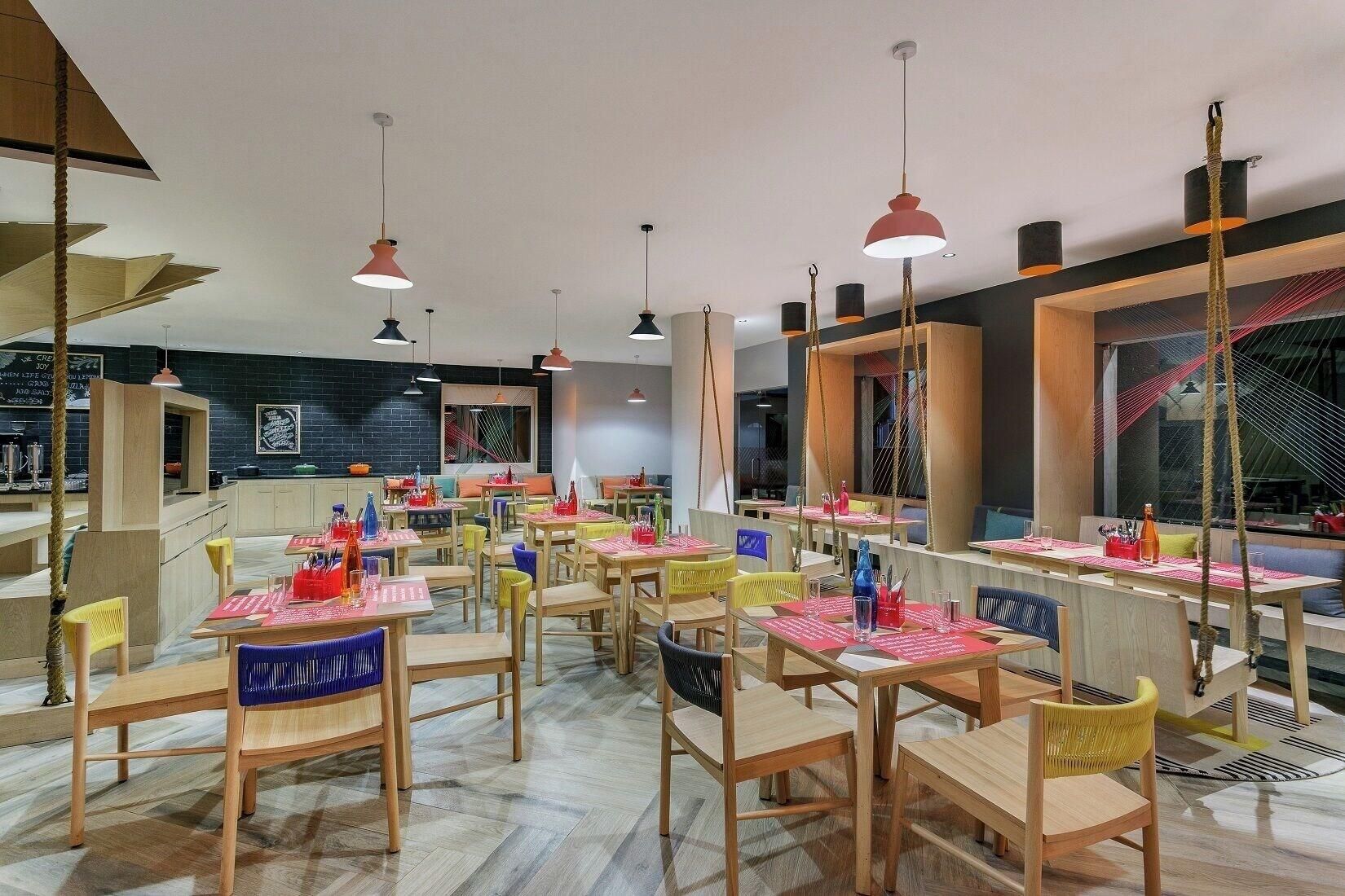 L’intérieur d’un restaurant moderne est agrémenté de tables et de chaises en bois, de guirlandes lumineuses suspendues et d’un mur en briques sur lequel est affiché un menu sur un tableau noir.