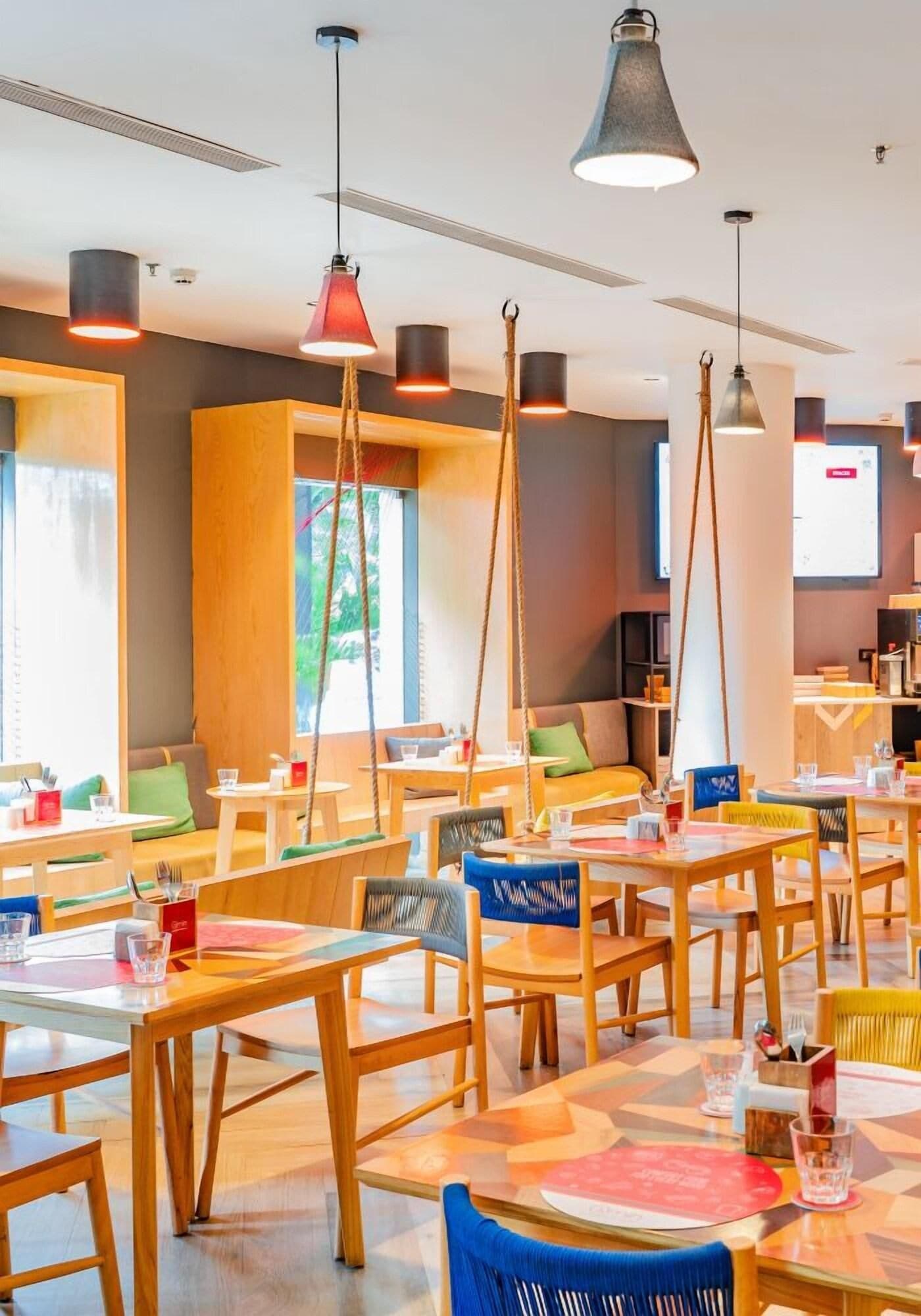 A modern restaurant with wooden tables and chairs, hanging pendant lights, and a view of the outdoors.