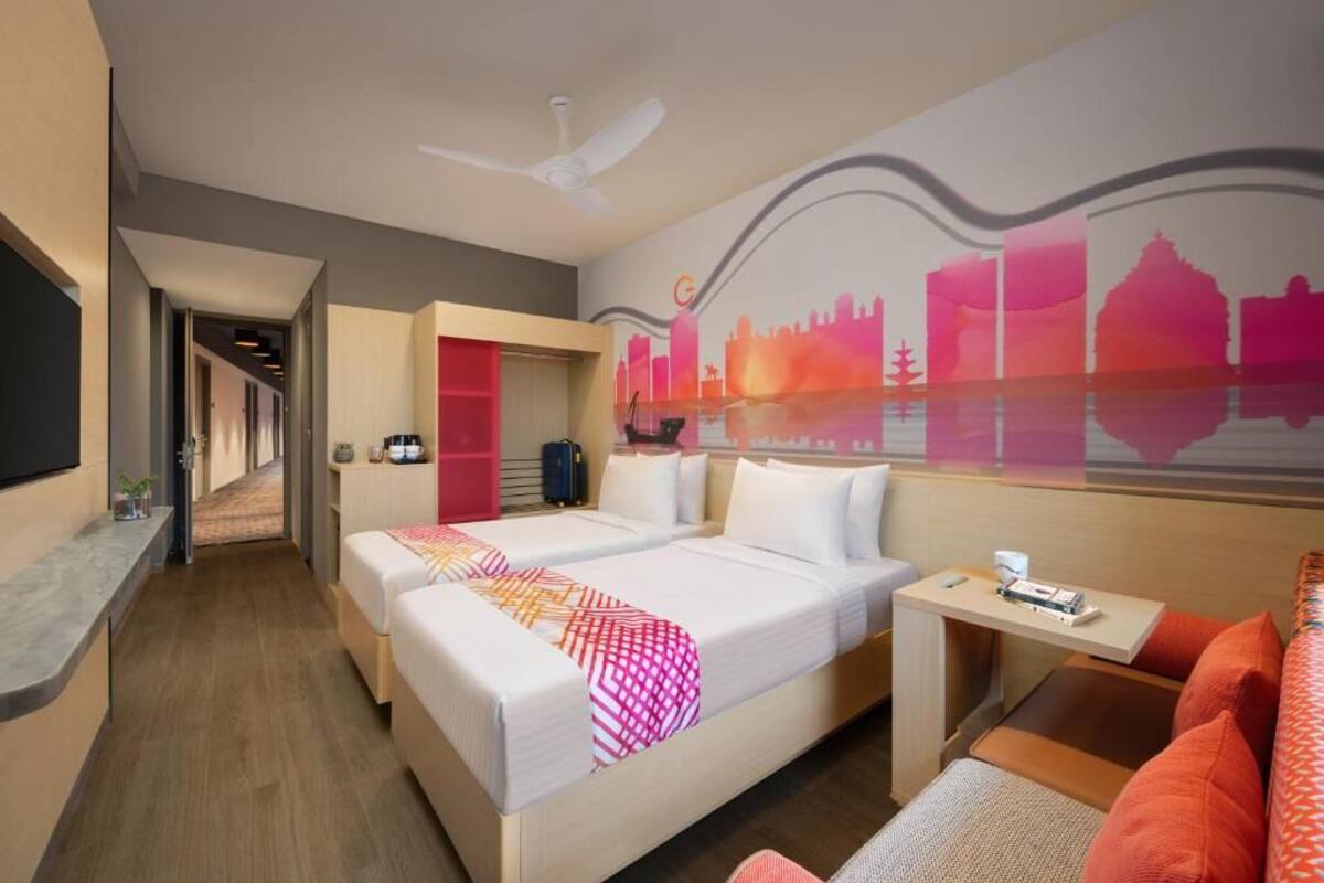 A hotel room with two beds, a desk, a chair, and a sofa. There is a cityscape mural on the wall and a ceiling fan.