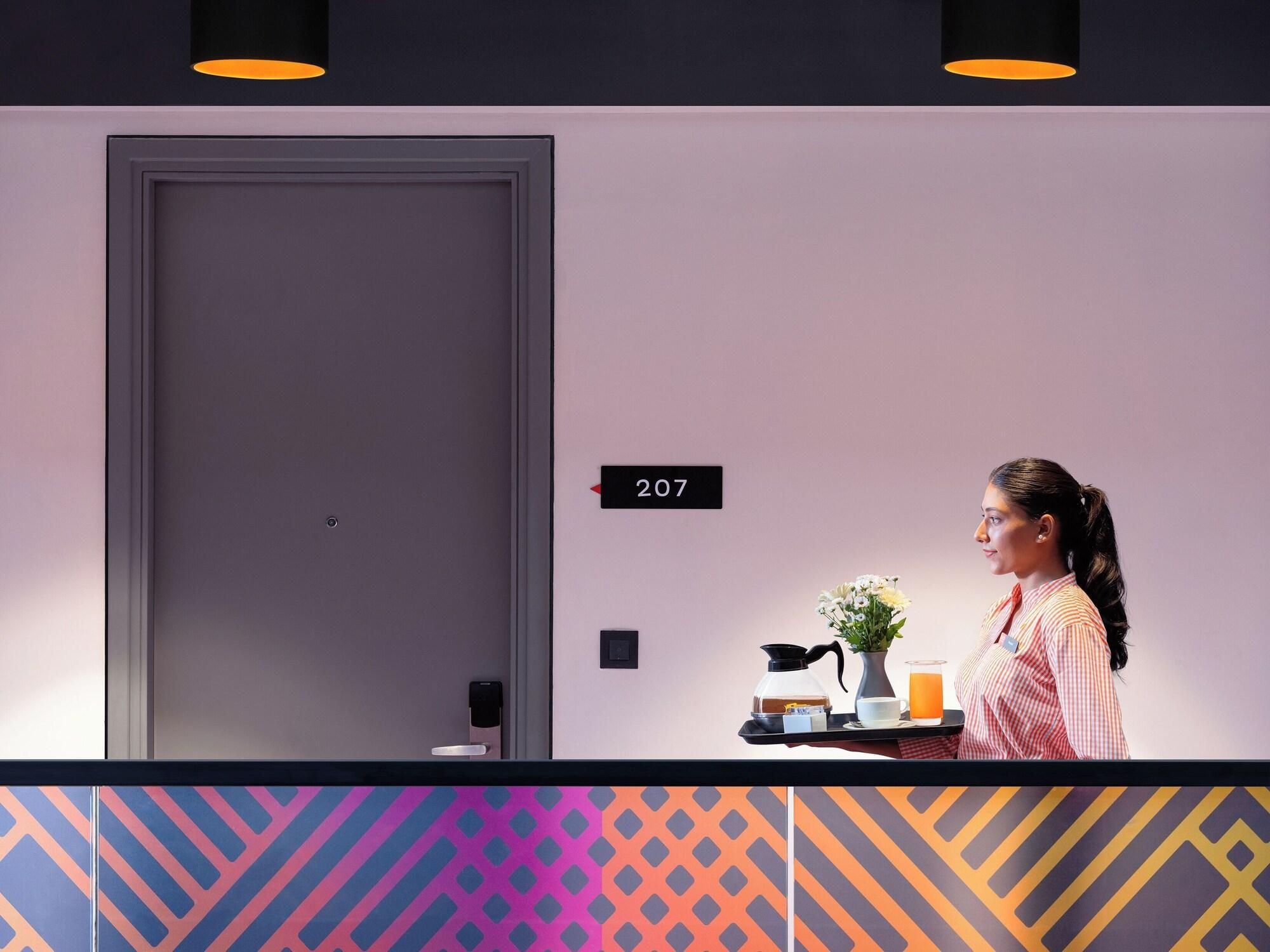 A hotel reception area with a closed door, a room number sign, a reception desk with a vase of flowers, a coffee maker, and a glass of orange juice.