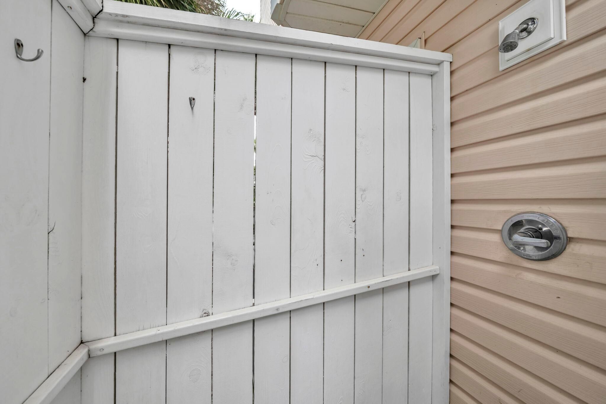 A white wooden fence with a metal gate, adjacent to a beige house with a window and a wall-mounted outdoor light fixture.