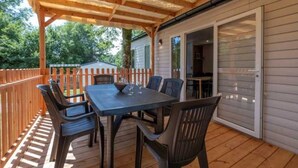 A covered patio with a table and chairs, a wooden deck, and a sliding glass door leading to an interior room. - ZEcamping 3* piscine chauffée hammam sauna mobil-home TV LV 49m2 (Verruyes)
