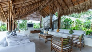 A modern living room with a thatched roof, white sofas, wooden chairs, and a staircase leading to an upper level. - Yeguas V60 by Wynwood House (Anapoima)