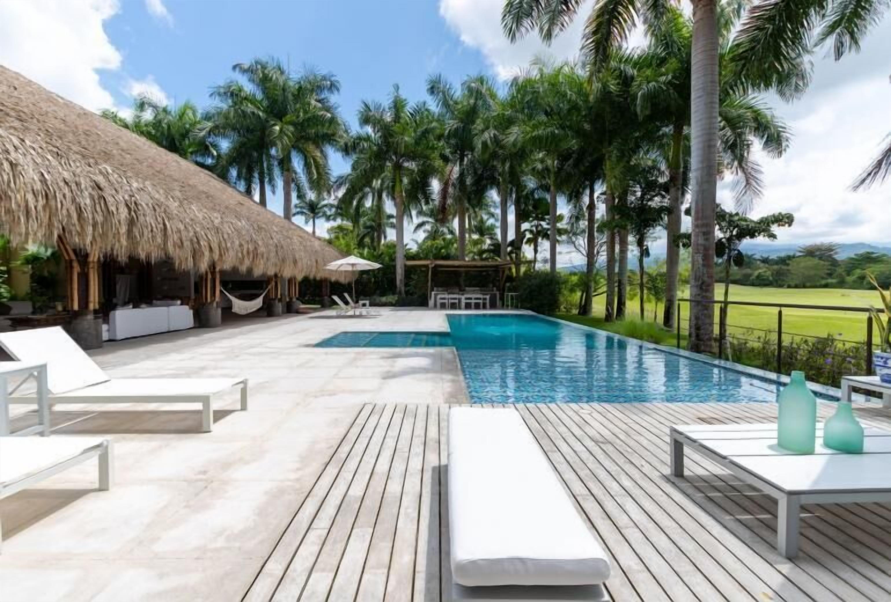 Zona de piscina con sillones de descanso, una estructura de techo de paja y vistas a palmeras y un paisaje verde.