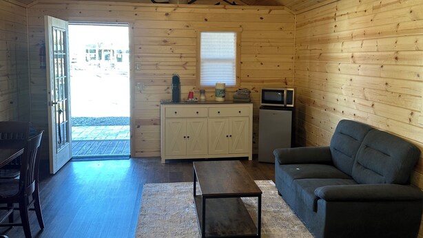 A cozy cabin interior with a wooden ceiling, a ceiling fan, a dining table with chairs, a sofa, a kitchen area with cabinets and a microwave, and a large window. - Cabin 3 with AC and WiFi in charming Reed Point. ADA compliant. (Reed Point)