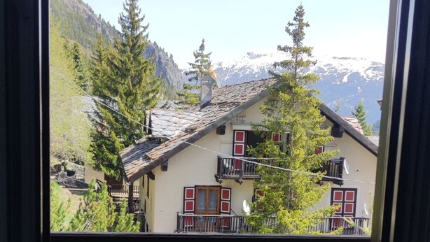 Balcón con piso de madera y baranda metálica, con vista a un edificio de detalles rojos y blancos y montañas al fondo.