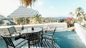 A terrace with a round table and chairs, a striped umbrella, and a view of the sea and hills.
