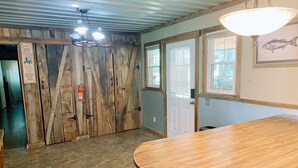 Ein Zimmer mit Holzboden, einem großen Holztisch und einem Deckenventilator. Es gibt zwei Türen und ein Fenster mit Jalousien.