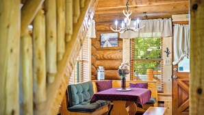 A cozy cabin interior with a wooden bench, a table with a purple tablecloth, a chandelier, and a window with curtains.