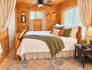 Une chambre dans une cabane en bois, avec un lit, des rideaux, un ventilateur de plafond et une table de chevet.