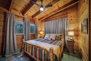 Ein Schlafzimmer einer Holzhütte mit Bett, Vorhängen, Deckenventilator, Nachttisch und Lampe.