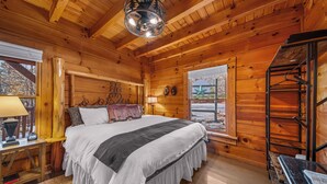 A cozy wooden cabin bedroom with a large bed, a nightstand with a lamp, a window with a view, and a wooden ceiling.