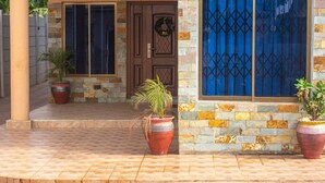 A building with a brown door, a blue window with a geometric pattern, and two potted plants on the ground. - Afranasa Inn (Tema)