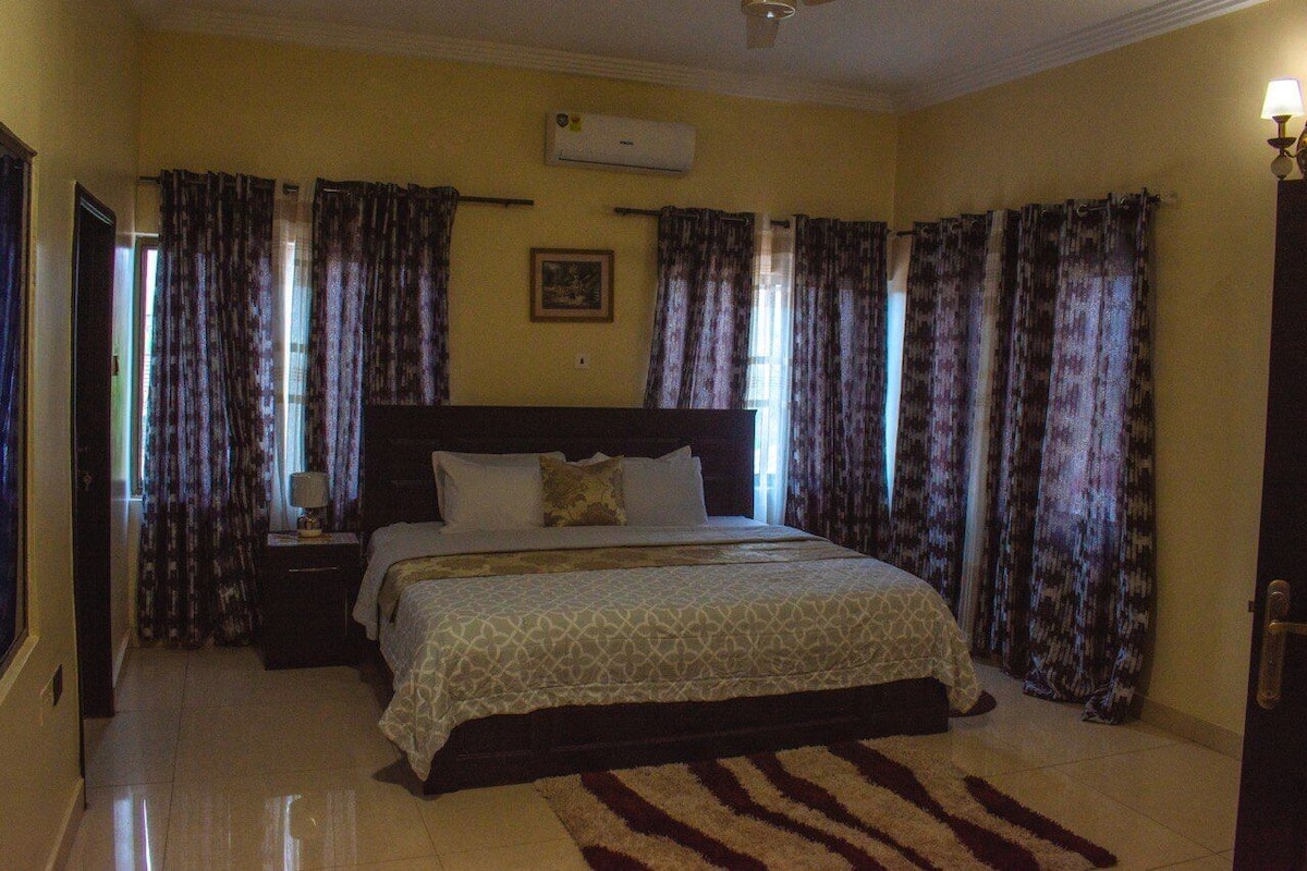 Quarto de hotel com uma cama grande, mesa de cabeceira, abajur e cortinas.