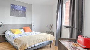 Een slaapkamer met een bed, een nachtkastje, een rode toaster en een raam met gordijnen.