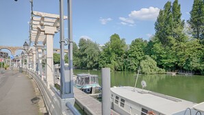 Een waterkantpad met een brug, een boot afgemeerd aan de steiger, en weelderige begroeiing langs de rivieroever.