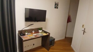 Een kleine kamer met een bureau, een aan de muur bevestigde televisie, een klein plantje en een rode handdoek die aan de deur hangt.